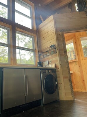 A kitchen with a stainless steel refrigerator, a washing machine, wooden cabinets, and a window with a view of trees. - Treehouse Escape w/Hot Tub, Near Smoky Mtn (Bryson City)