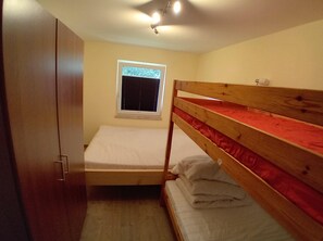 Ein kleines Zimmer mit einem Etagenbett, einem Kleiderschrank, einem Fenster mit Jalousien und einer Deckenleuchte.
