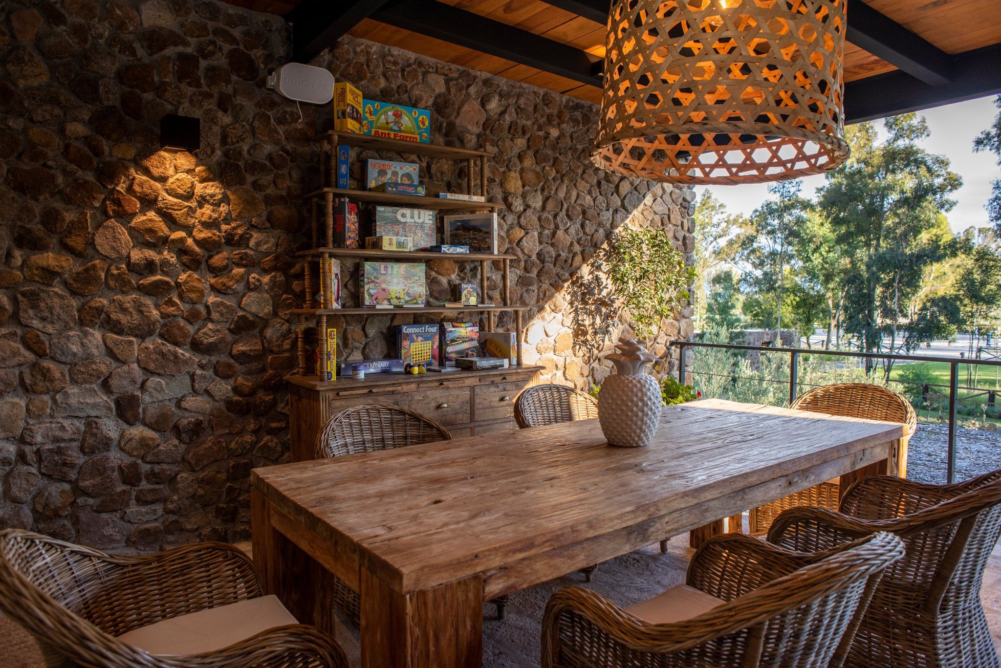 Un comedor al aire libre rústico con pared de piedra, mesa de madera, sillas de mimbre y una estantería llena de juegos de mesa.
