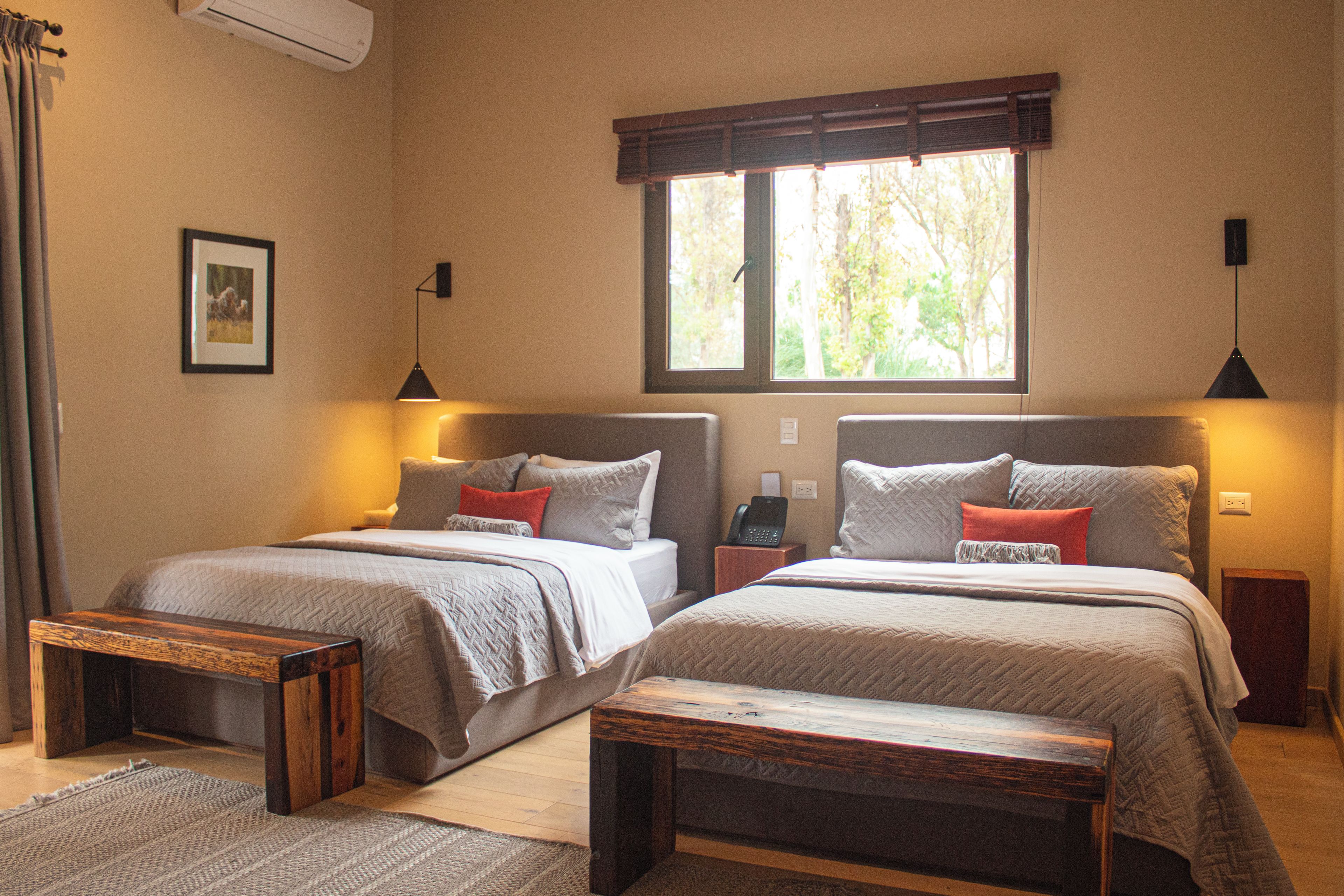 A double room with two beds, a small table, a bedside table with a phone, and a window with blinds.