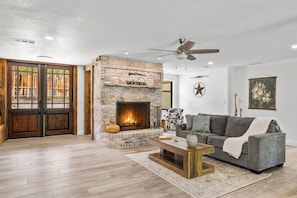 A living room with a stone fireplace, a grey sofa, a coffee table, and a ceiling fan. - The Castle Creek Ranch - 5-Acre Ranch, Private Pool, Game Room & Country Getaway (Cross Roads)
