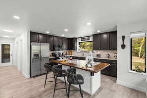 A modern kitchen with dark cabinets, stainless steel appliances, a marble countertop, and a large window offering a view of trees.