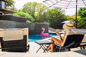 Uma pessoa relaxando em uma espreguiçadeira à beira da piscina, piscina com boia de flamingo e um muro de pedra ao fundo.