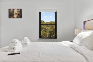 Quarto de hotel com cama, roupa de cama branca, controle remoto, janela com vista para árvores e um quadro na parede.