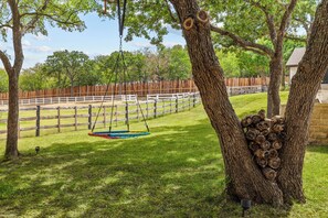 A fenced outdoor area with a swing set, trees, and a pile of logs. - The Castle Creek Ranch - 5-Acre Ranch, Private Pool, Game Room & Country Getaway (Cross Roads)