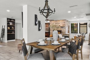 Uma sala de jantar com uma mesa de madeira posta para quatro pessoas, rodeada por cadeiras com encostos de palha. Um candelabro pendurado paira acima, e uma lareira de pedra é visível ao fundo.