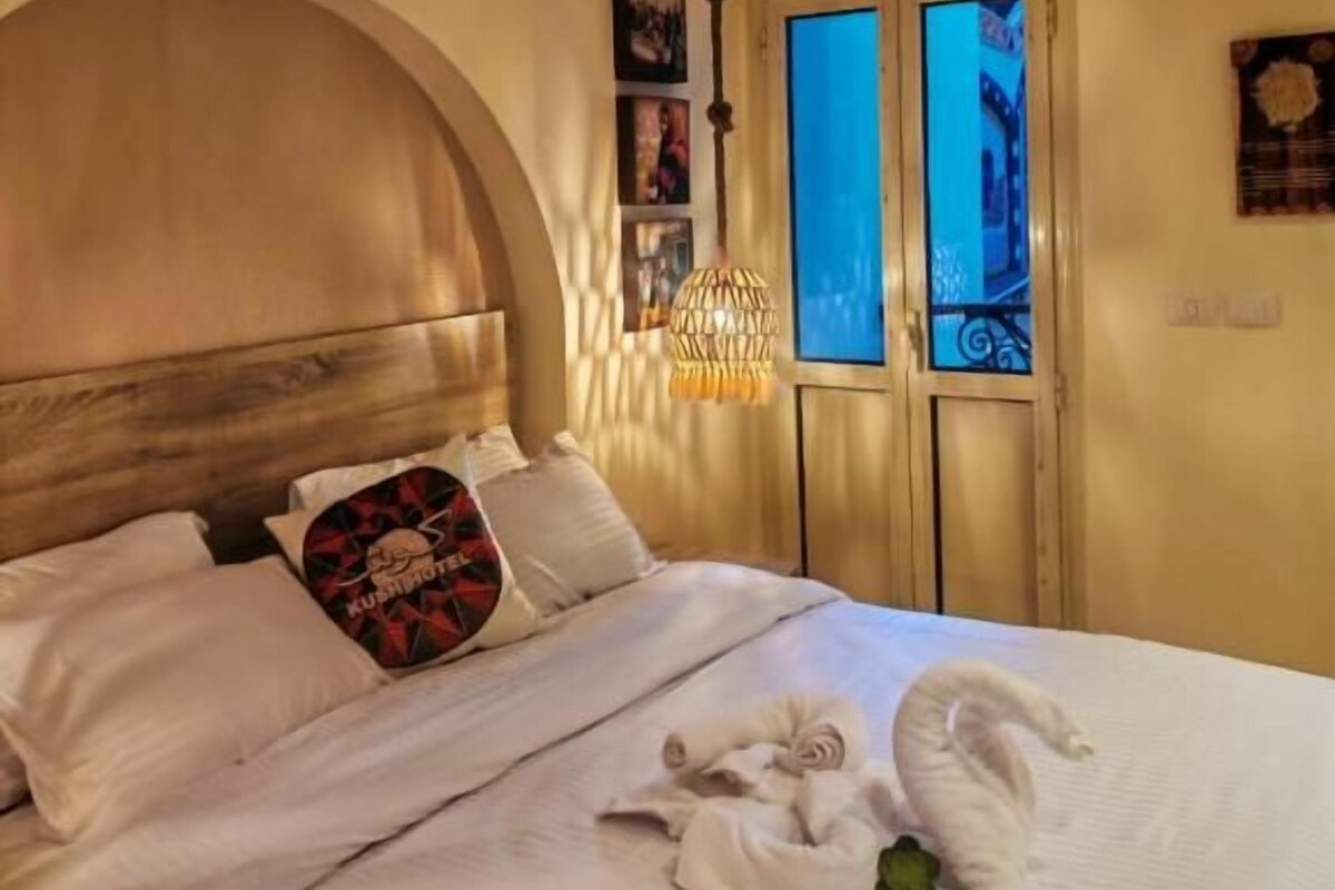 Una habitació d'hotel amb un llit gran, un panell de fusta al capçal, una finestra amb cortines blaves i un aire condicionat fixat a la paret.