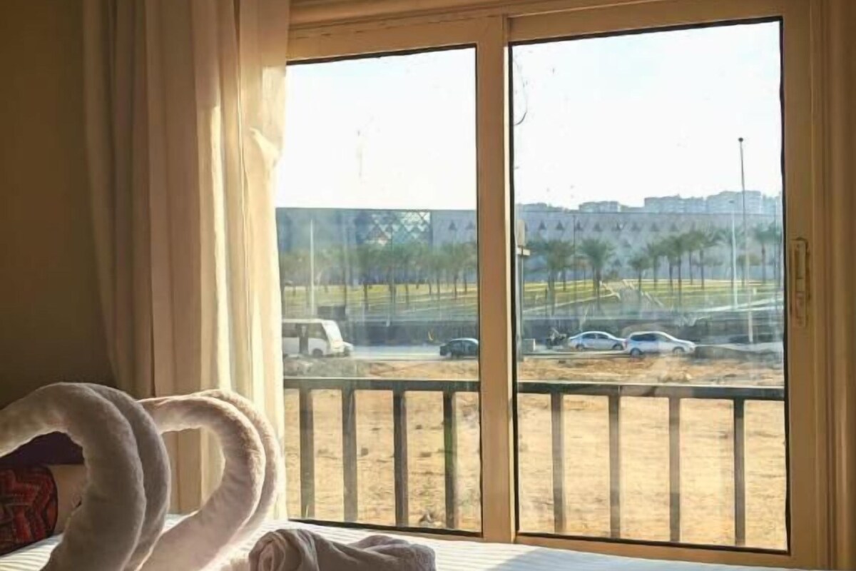 Habitació d'hotel amb un llit, una finestra gran que ofereix vista a una zona d'aparcament i edificis, i una tovallola amb forma de cigne sobre el llit.