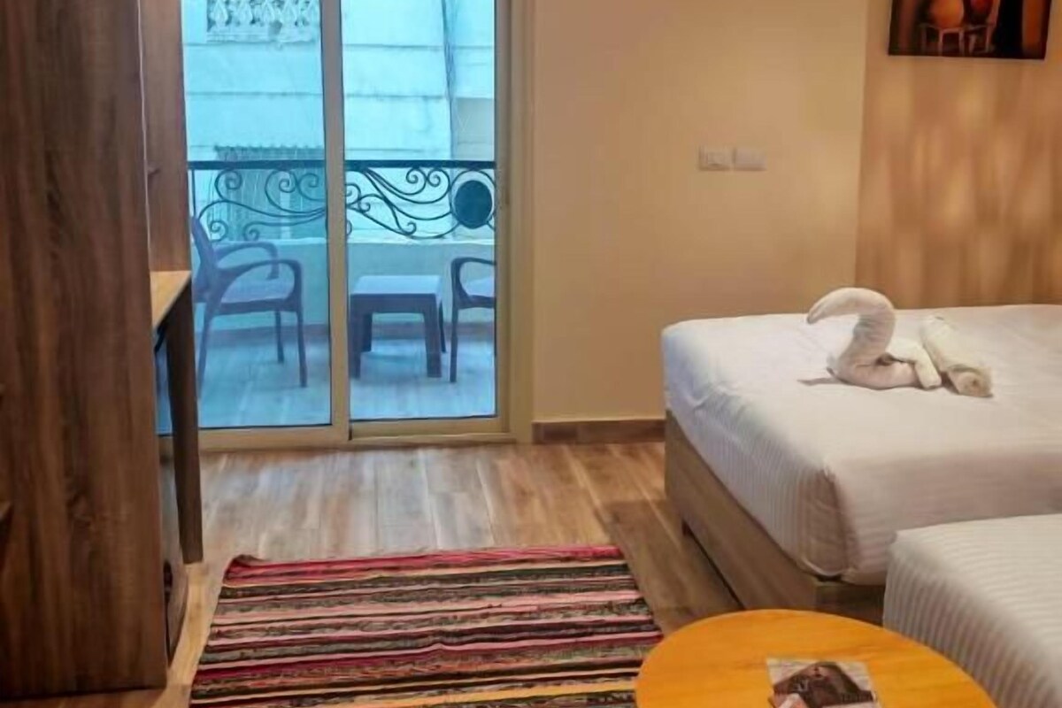 Una habitació d'hotel amb un llit, una taula rodona petita, una estora amb ratlles de colors, un balcó amb cadires i un aparell d'aire condicionat.