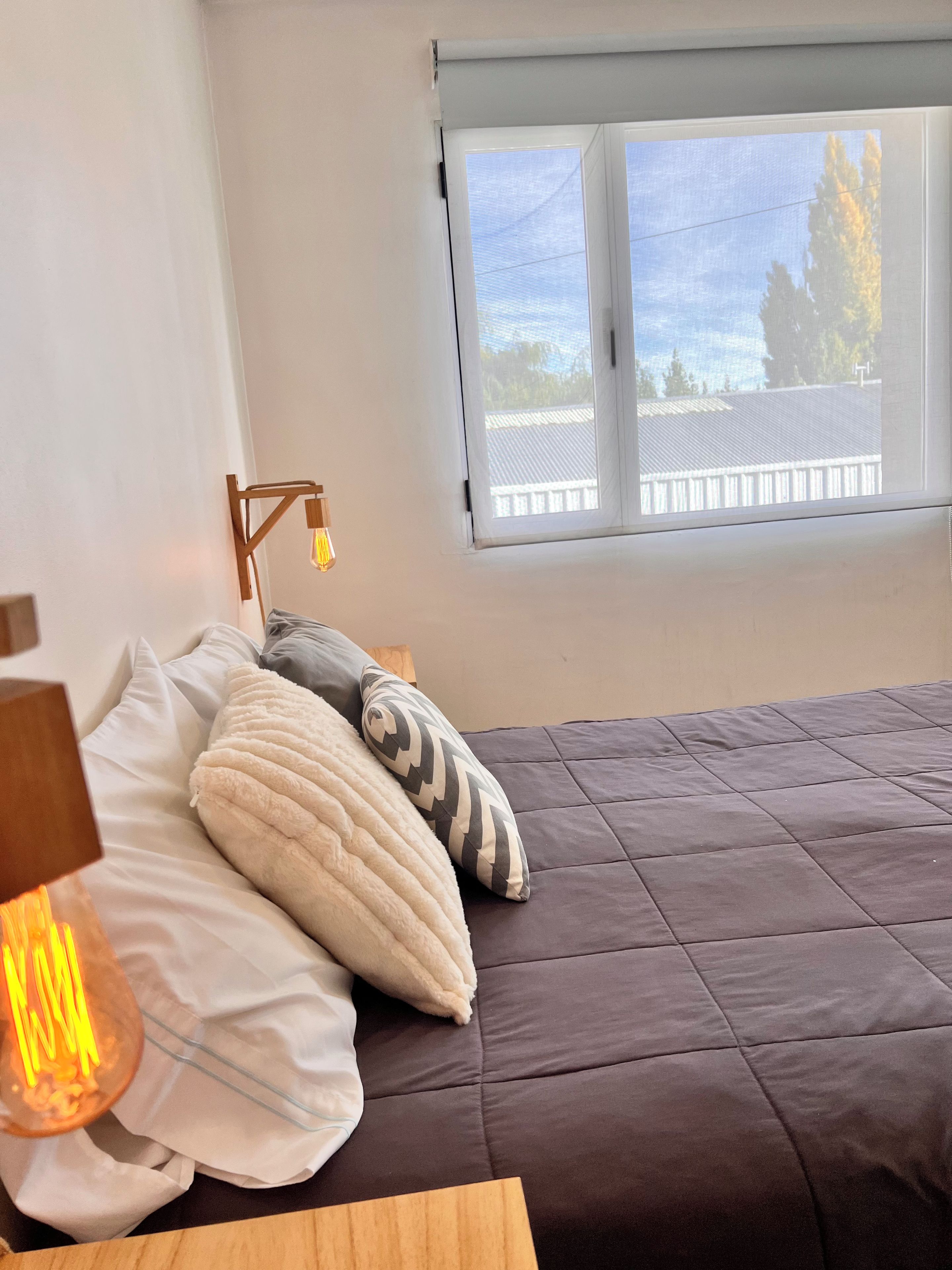 A bedroom with a bed, pillows, a bedside table with a lamp, and a window with a view of a fence and trees.