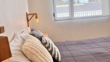 A bedroom with a bed, pillows, a bedside table with a lamp, and a window with a view of a fence and trees.