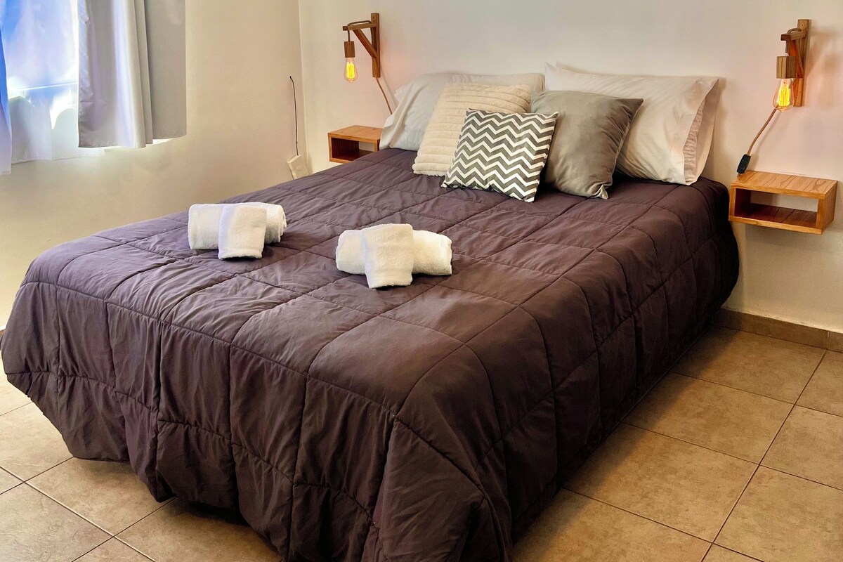 A bed with a dark-colored duvet, two pillows, and two folded towels. There are two bedside tables with wall-mounted lamps.