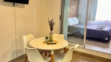 A room with a round table, white chairs, a flat-screen TV mounted on the wall, and a sliding glass door leading to a bed.