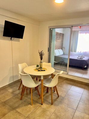 Una habitación con una mesa redonda, sillas blancas, un televisor de pantalla plana en la pared y un ventanal corredero que da a la cama.
