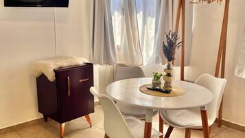 A modern living room with a round dining table, white chairs, a dark-colored cabinet, a flat-screen TV mounted on the wall, and a window with sheer curtains.