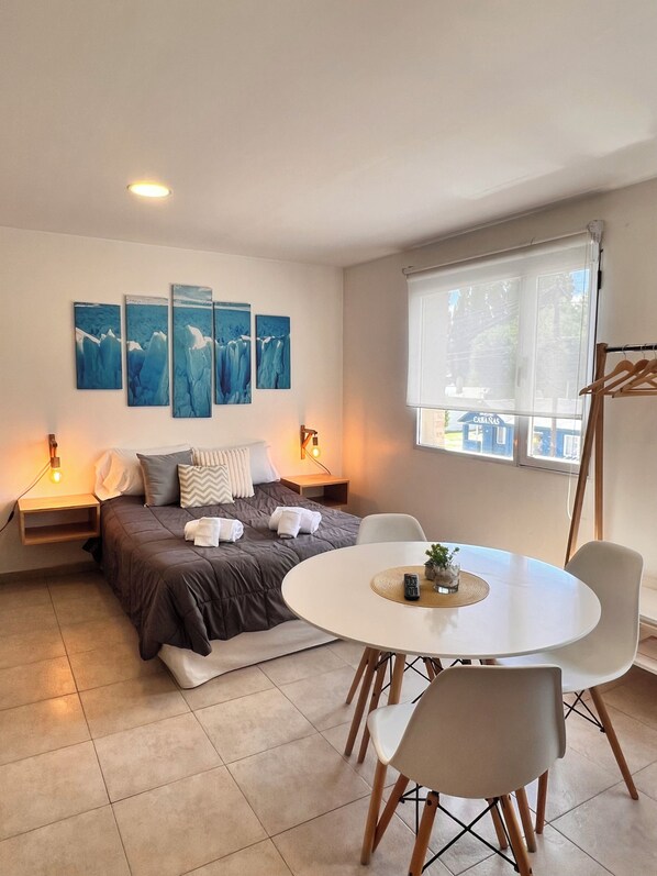 A modern bedroom with a bed, bedside tables, a round dining table with chairs, and a window with a view.