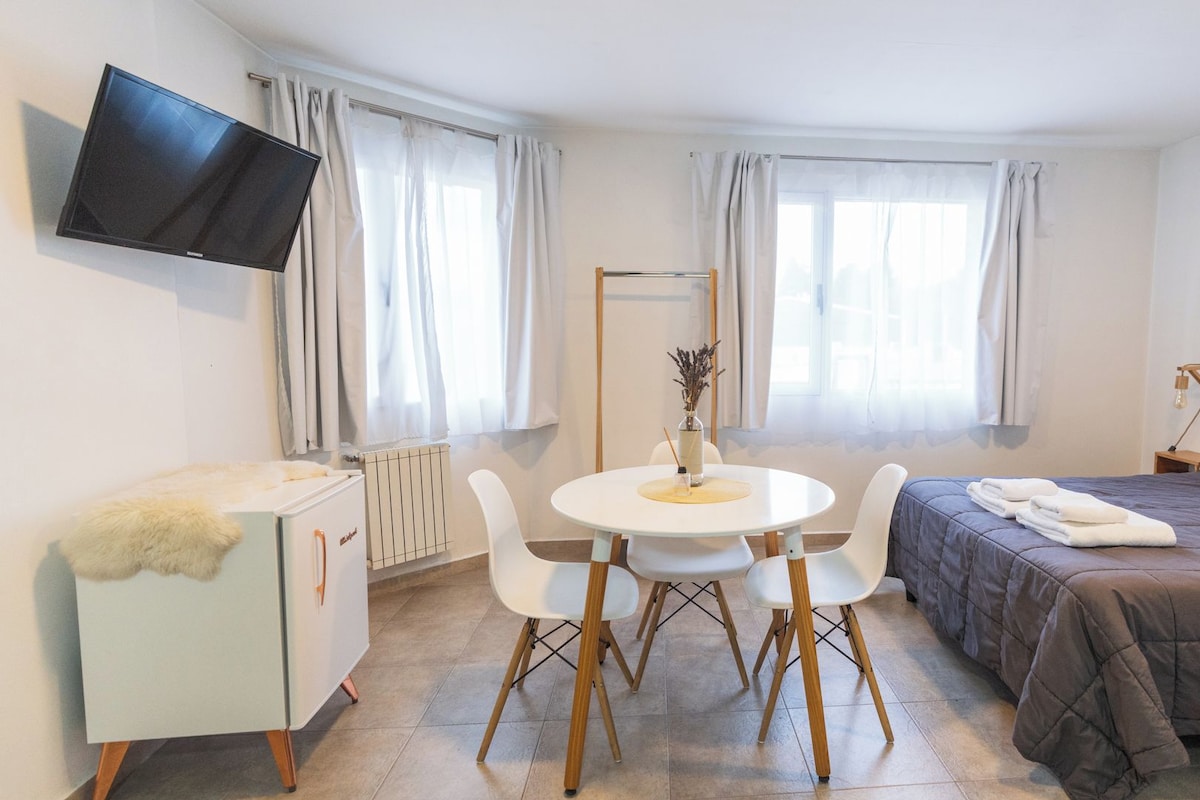 A hotel room with a bed, a desk, a chair, a TV mounted on the wall, and a window with curtains.