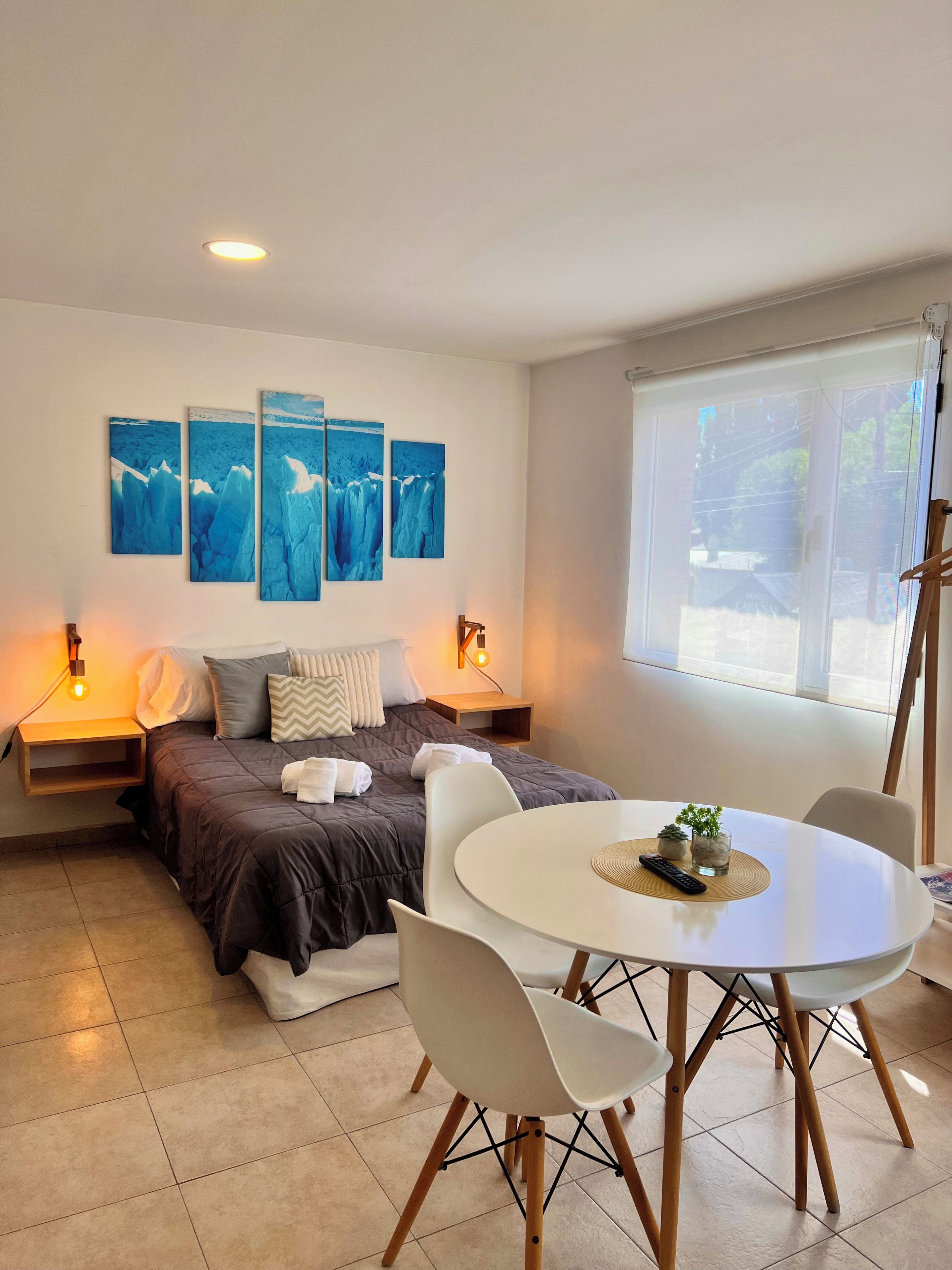 Quarto moderno com cama, mesas de cabeceira, arte na parede, uma janela e um espaço de jantar com cadeiras e mesa.