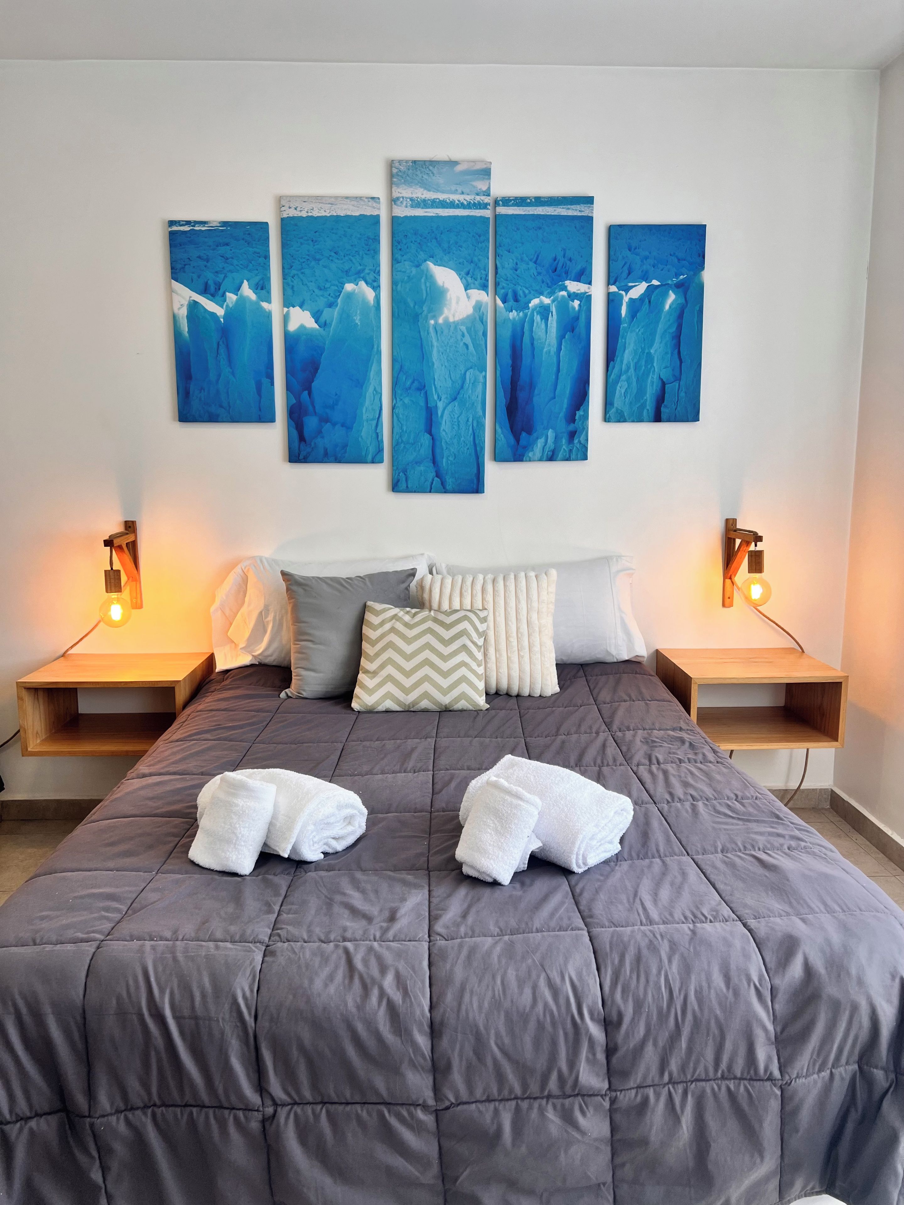 Quarto com uma cama grande, duas mesas de cabeceira, luminária fixada na parede e uma obra de arte com vários painéis sobre a cama.
