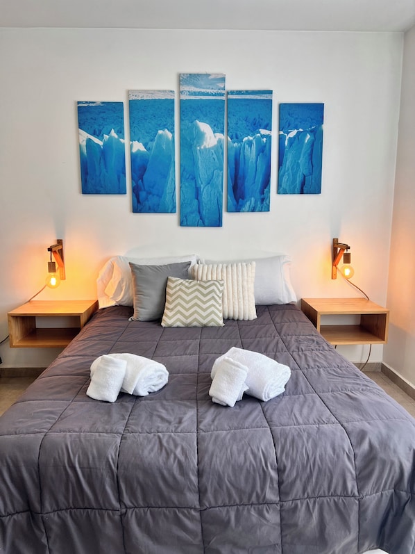 A bedroom with a large bed, two bedside tables, a wall-mounted light fixture, and a multi-panel artwork above the bed. - Espora (El Calafate)