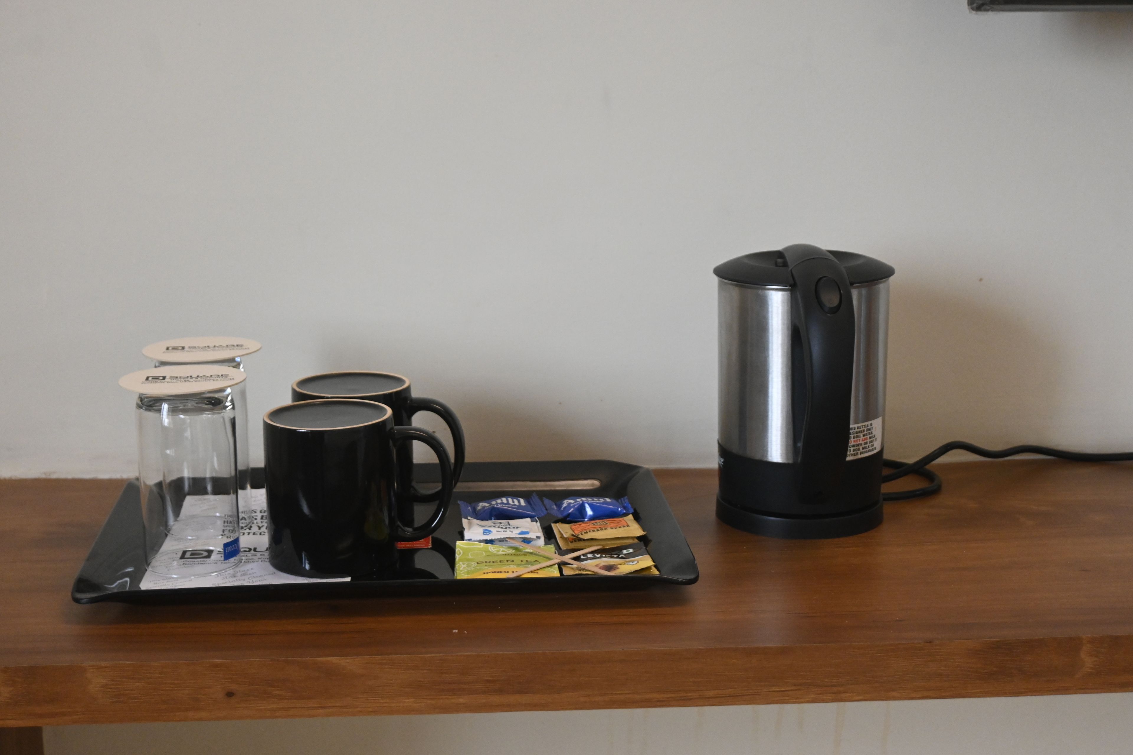A tray with a coffee maker, a glass carafe, two black mugs, and several packets of tea on a wooden table.