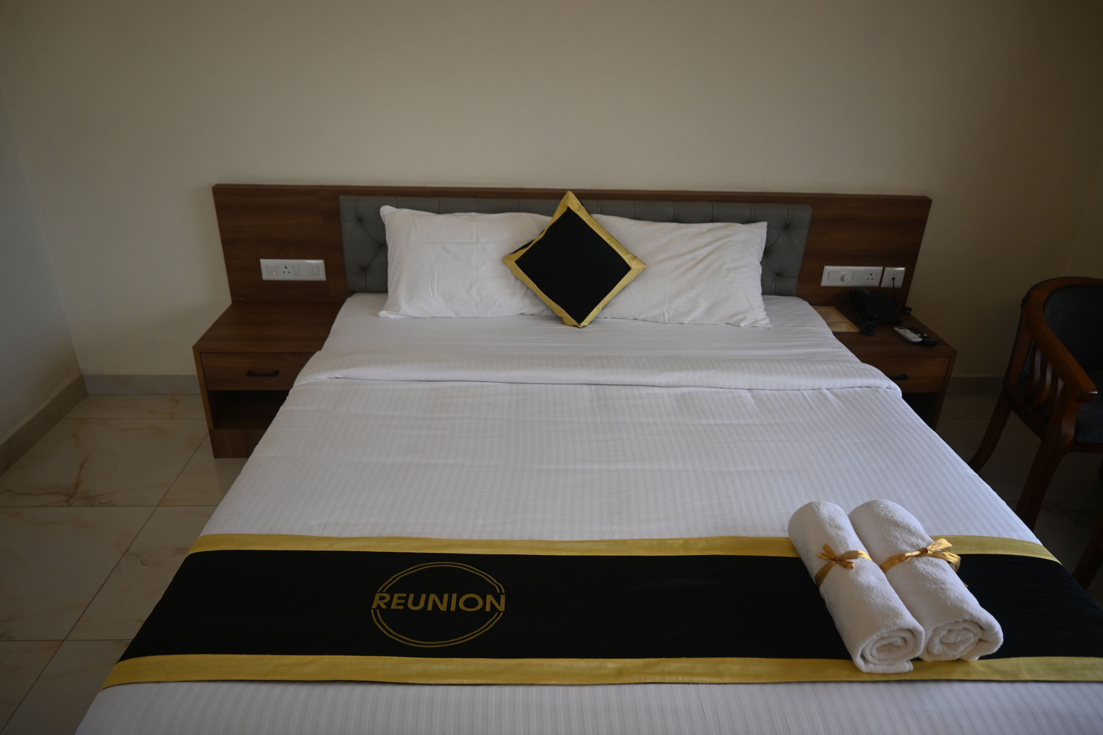 A hotel room with a neatly made bed, a bedside table, a chair, and a wall socket.