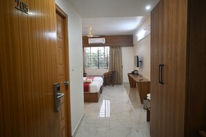 A hotel room with a wooden door, a bed, a desk, a chair, a TV, and a window with curtains.