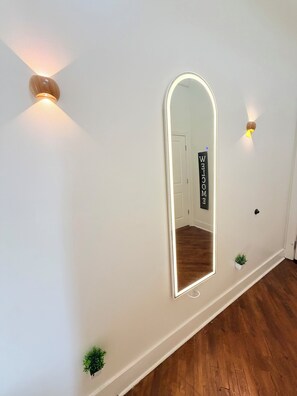 A hallway with a large mirror, wall-mounted lights, a welcome sign, and small potted plants.