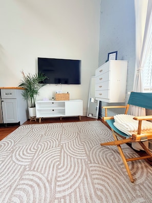 A modern living room with a flat-screen TV, a white cabinet, a wooden chair, a patterned rug, and a potted plant.