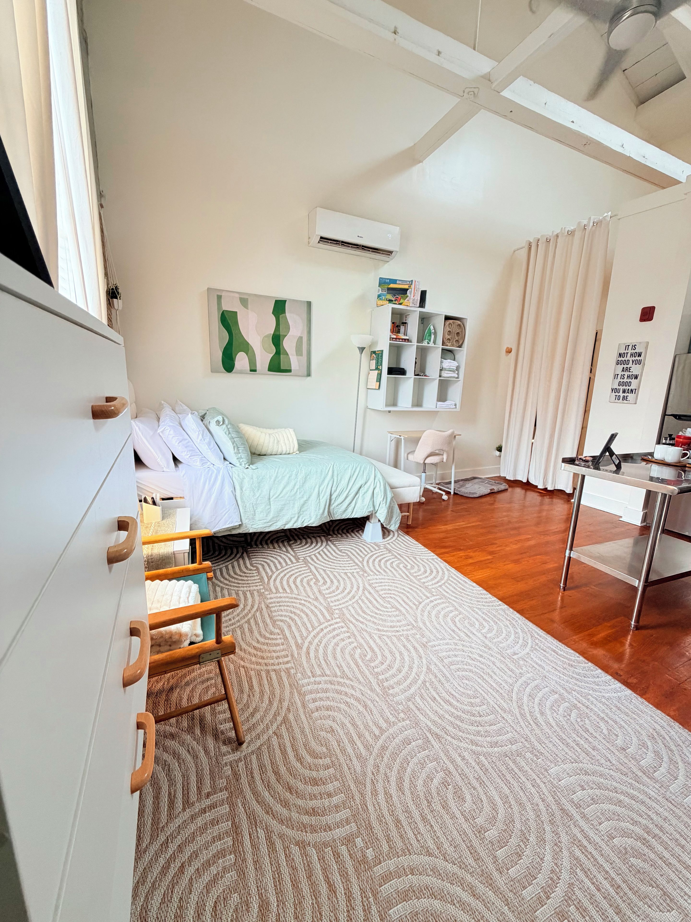 A bedroom with a bed, a desk, a chair, a wardrobe, a radiator, and a wall with a motivational quote.