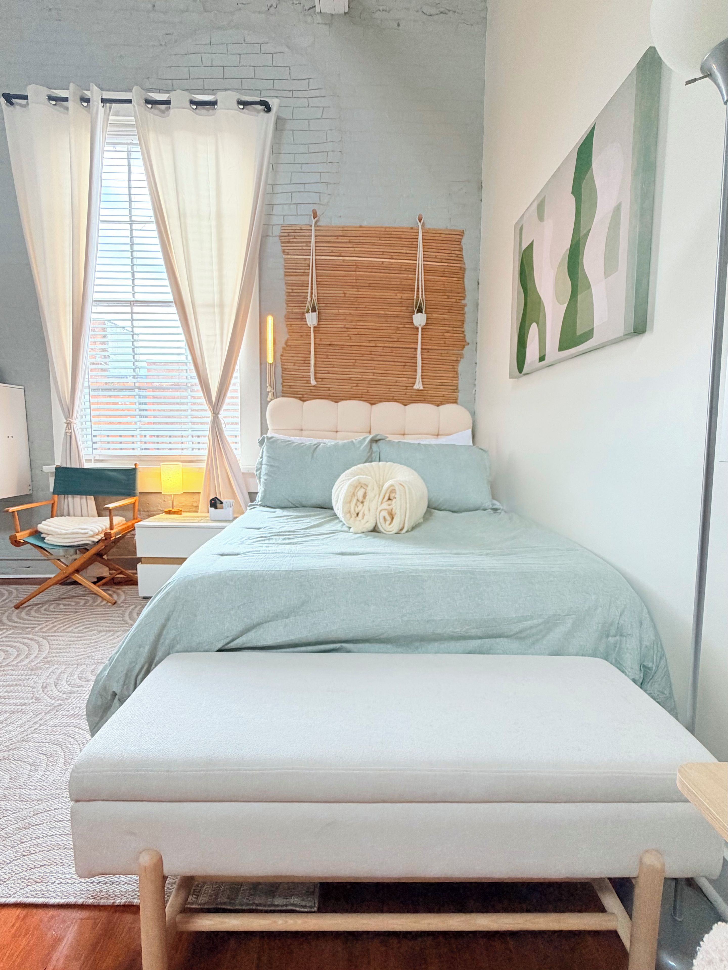 Une chambre à coucher avec un lit, une chaise, une table de chevet, une lampe et une fenêtre avec des rideaux.