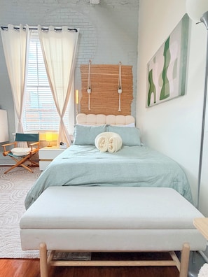 Un dormitorio con cama, una silla, mesita de noche, lámpara y ventana con cortinas.