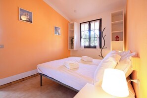 Ein Schlafzimmer mit Bett, einem Fenster mit Vorhängen, einem Nachttisch mit Lampe und zwei Wandregalen.
