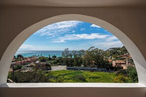 Blick auf eine Küstenstadt durch ein Rundbogenfenster, mit Gebäuden, Grünflächen und dem Meer.