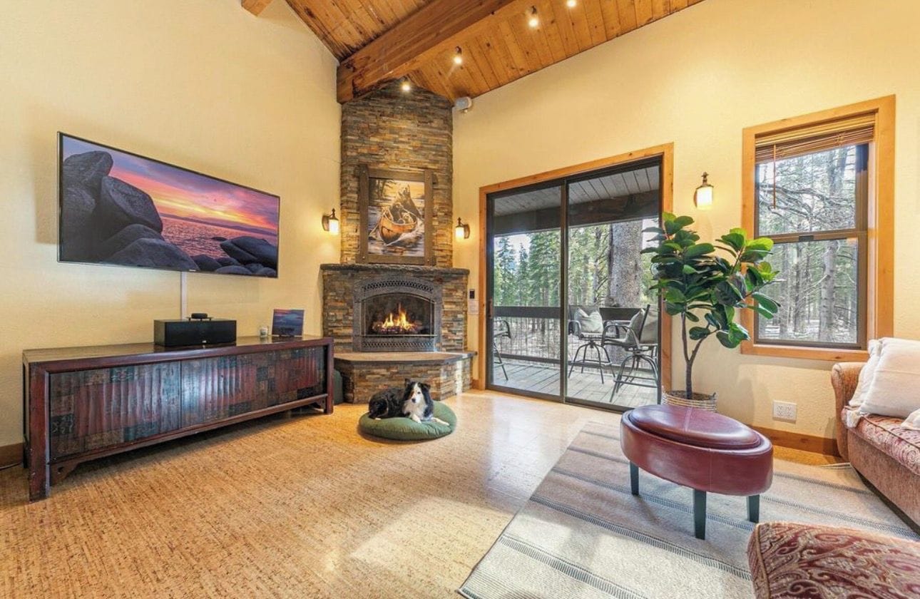 A living room with a fireplace, a television mounted on the wall, a dog bed, and a large window overlooking a deck.