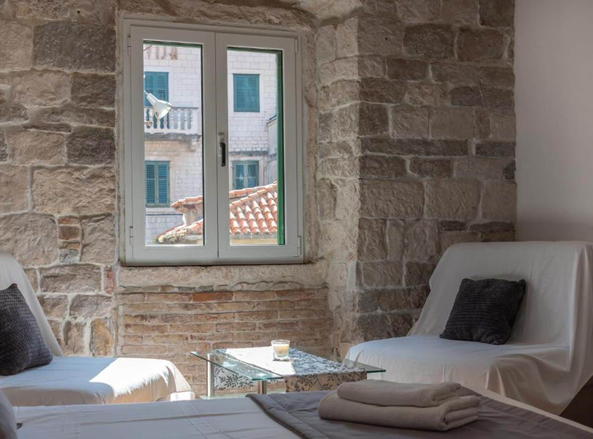 A room with stone walls, a window with shutters, two white chairs, a small table with a glass, and a bed with a white cover.