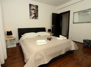 Un letto rifatto con copriletto, due comodini con lampade, un quadro appeso al muro e una porta sulla destra.