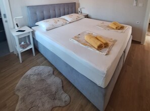 Ein Bett mit grauem Kopfteil, ein Nachttisch mit Lampe und ein aufgerolltes Handtuch auf dem Bett.