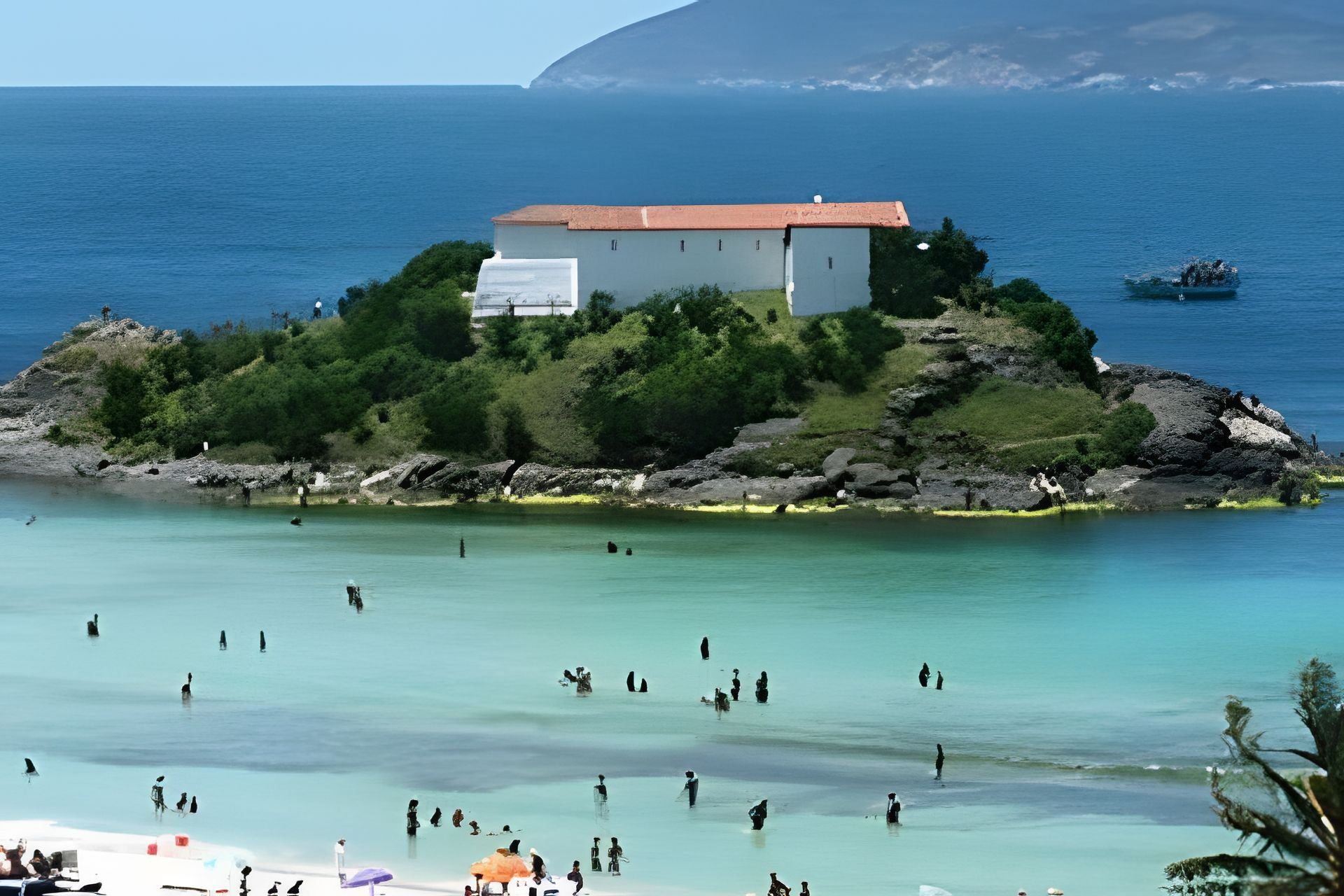 A coastal building surrounded by greenery, overlooking a clear blue sea with people in the water.