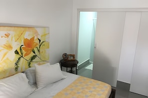 A bedroom with a bed, a nightstand, a painting, and a door leading to another room. - Casa Praia das Dunas (Cabo Frio)
