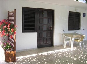 A white building with a dark wooden door and shutters, a small table with chairs, and a wooden lattice structure with flowers. - Three Bedroom Apartment with Terrace (A2) (Marina)
