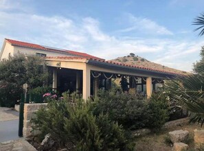 A building with a red-tiled roof, a covered patio area, and a landscaped garden with shrubs and a palm tree. - Five Bedroom Apartment with Sea View (National Park Kornati)