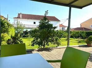 A covered outdoor area with a table and chairs, a small tree, and a building with a red-tiled roof in the background. - Two Bedroom Apartment with Balcony and Garden View (Trogir)