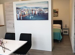 A hotel room with a bed, a nightstand, a painting on the wall, and a dining area with chairs and a table. - Two Bedroom Apartment with Balcony and Garden View (Trogir)