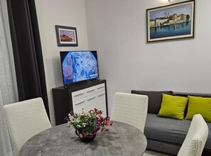 Una habitación con una televisión sobre un mueble bajo, un sofá con cojines, una mesa de comedor con un florero y dos sillas.