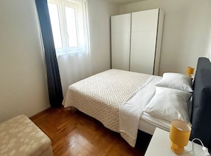 A hotel room with a bed, a bedside table with a lamp, a window with curtains, and a wooden floor. - One Bedroom Apartment with Balcony and City View 2 (Trogir)