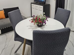 A modern living room with a round dining table, grey patterned chairs, a sofa with cushions, a coffee table with flowers, and a TV on a cabinet. - One Bedroom Apartment with Pool 2 (Trogir)