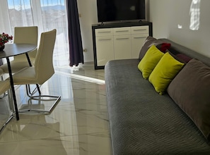 A living room with a sofa, a TV, a dining table with chairs, and a view of the outside. - One Bedroom Apartment with Garden View and Pool (Trogir)