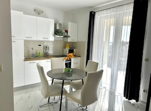 A modern kitchen with white cabinets, a dining table with chairs, and a view of the city through large windows. - One Bedroom Apartment with Garden View and Pool (Trogir)