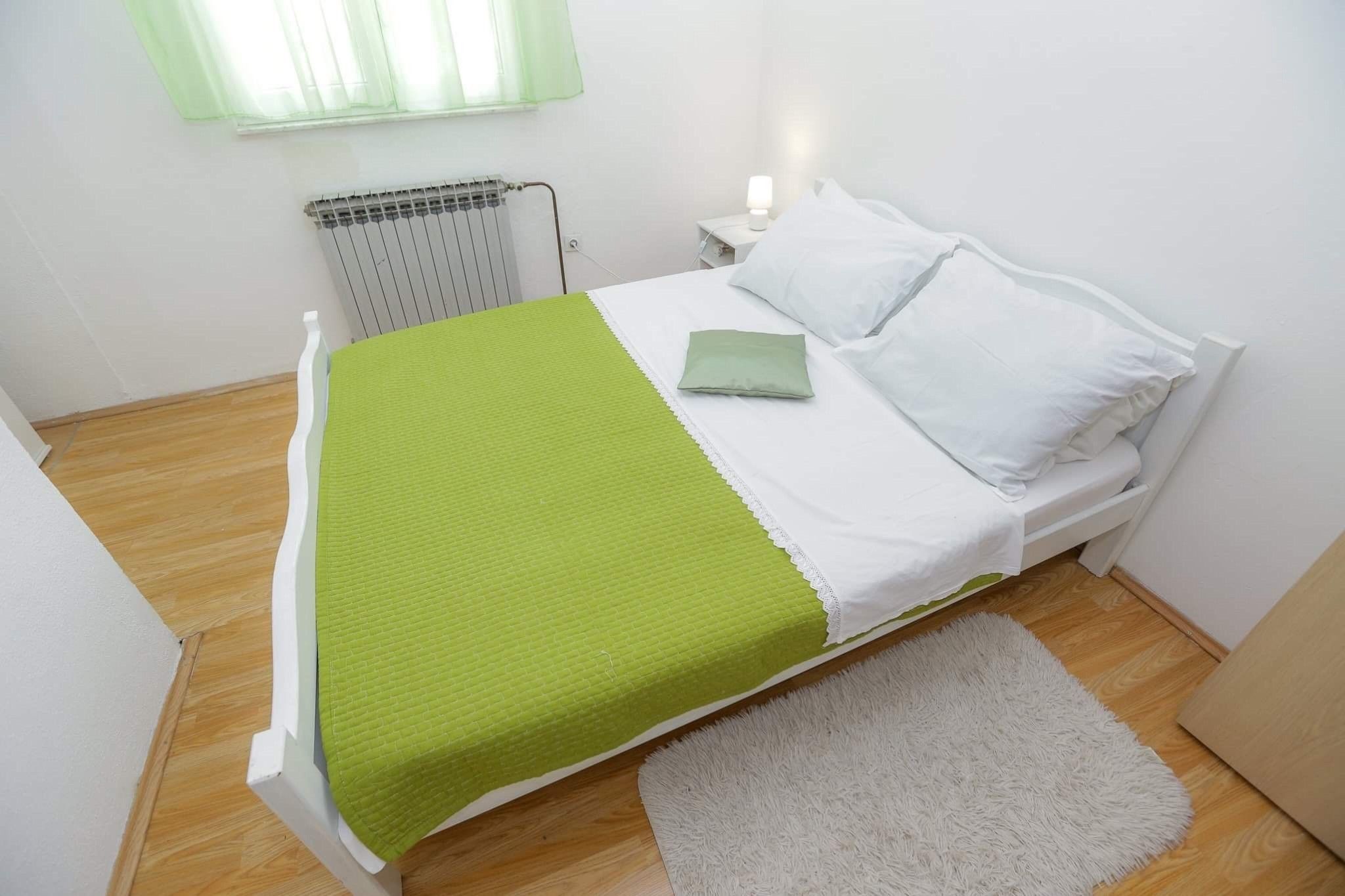 Una cama con colchón blanco y ropa de cama verde, una mesita de noche blanca con lámpara, un radiador y una alfombra blanca sobre piso de madera.
